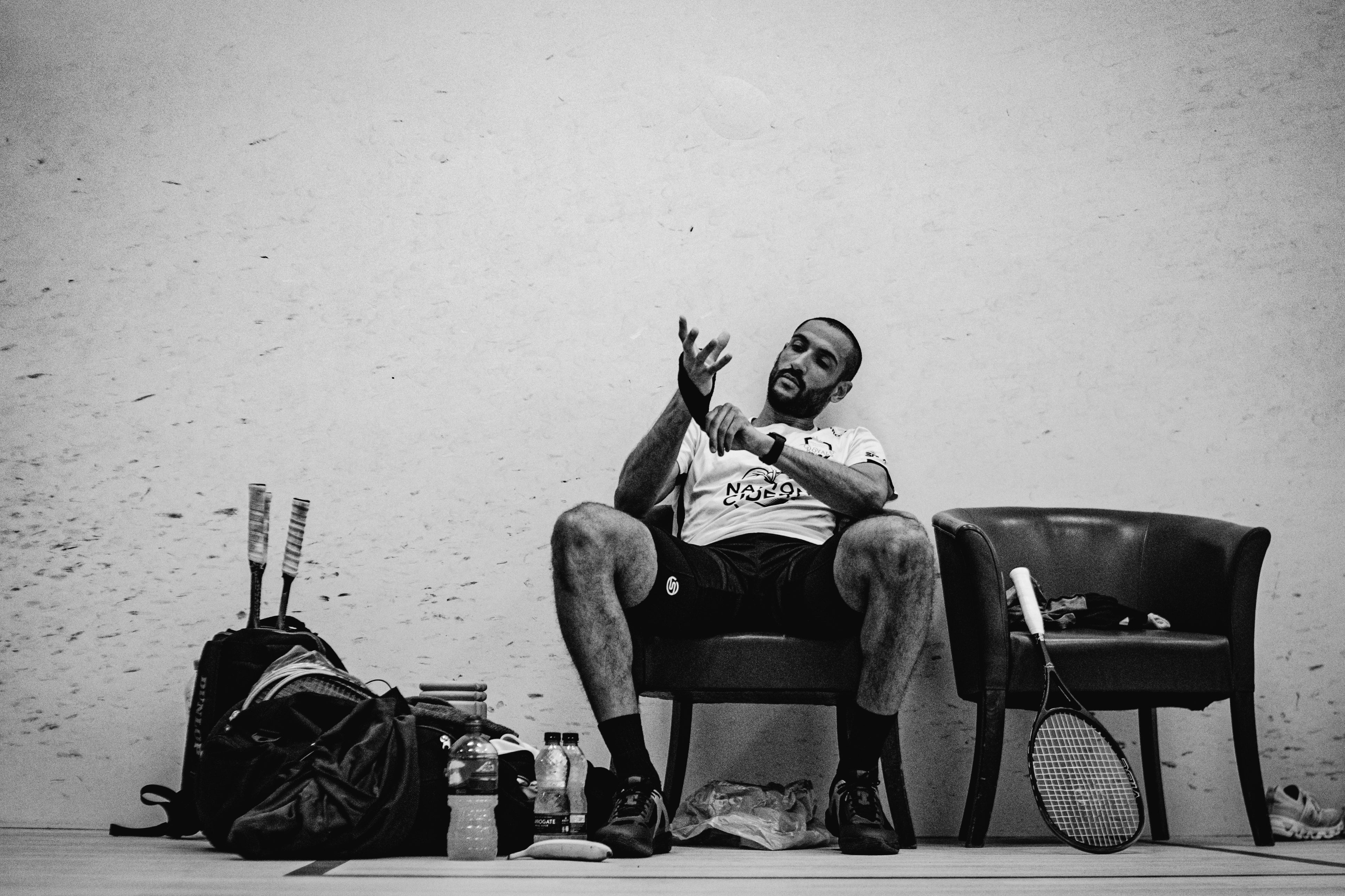 Declan James sitting on a chair with squash equipment around him, black and white photo
